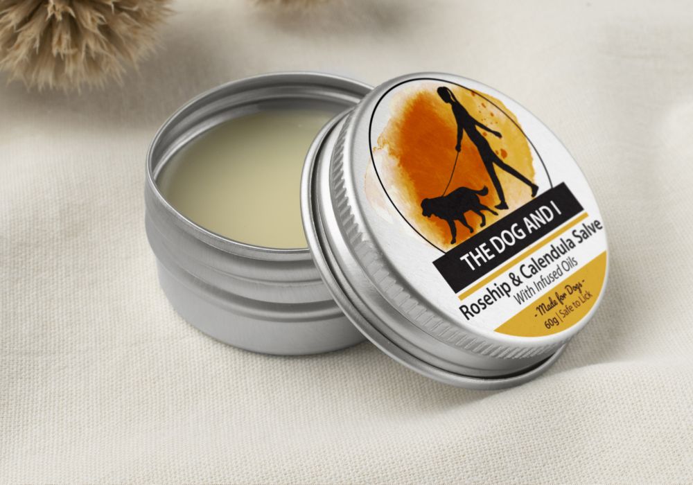 A tin container with a label showing a silhouette of a dog and a person with a dog leash, indicating the product is for pets. The tin is open, revealing a yellowish salve inside. The brnading is The Dog and I